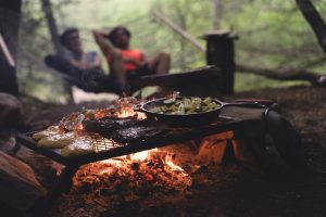 A picture of people camping and cooking around a campfire. In focus is the campfire with a grill over with a pan of cucumbers and onion and on the grill is various meats and chicken.