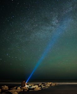 A person sitting near some rocks and shining their flashlight in the sky. You can see a long beam coming from the flashlight.