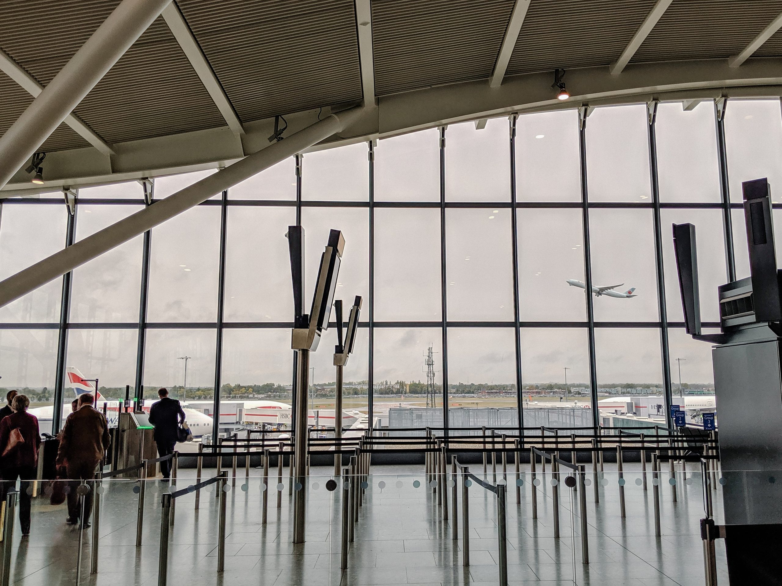 A picture of an airport security line with a few people in it who are traveling. The wall of the room is glass so that the viewer can see a plane take off in the background