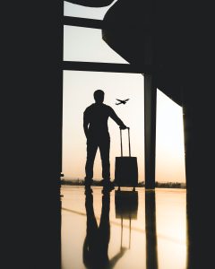 A silhouetted man and his suitcase stand near a window as a plane is taking off in the background.