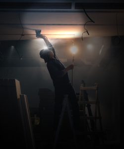A person leans back on a ladder to fix something that is on the ceiling. They wear a headlamp to help illuminate the area they are working.